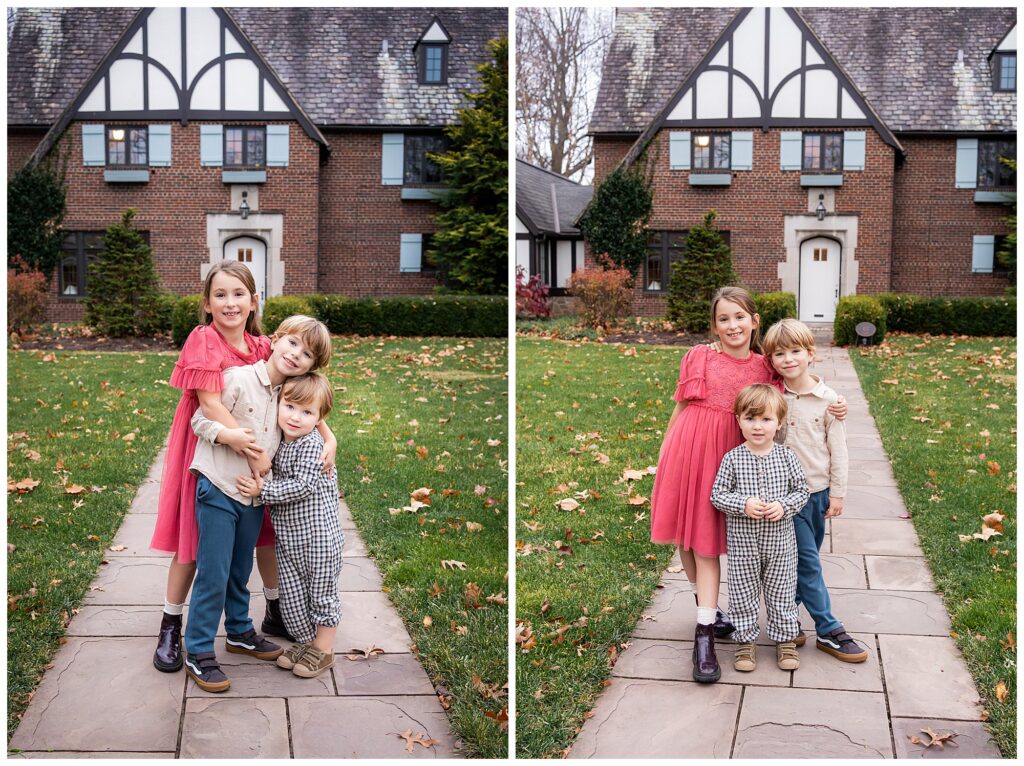 Three children pose and smile together on a stone pathway in front of a large brick house with Tudor-style architecture.