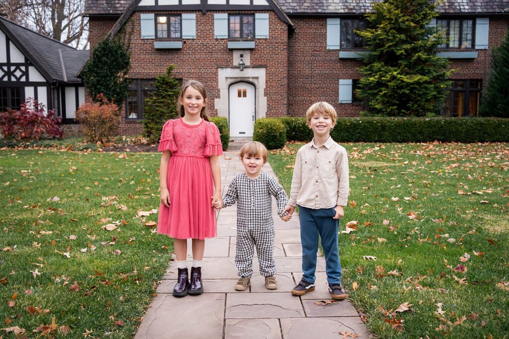 Three young children stand on a stone path in front of a brick house with Tudor-style details, holding hands and smiling at the camera.