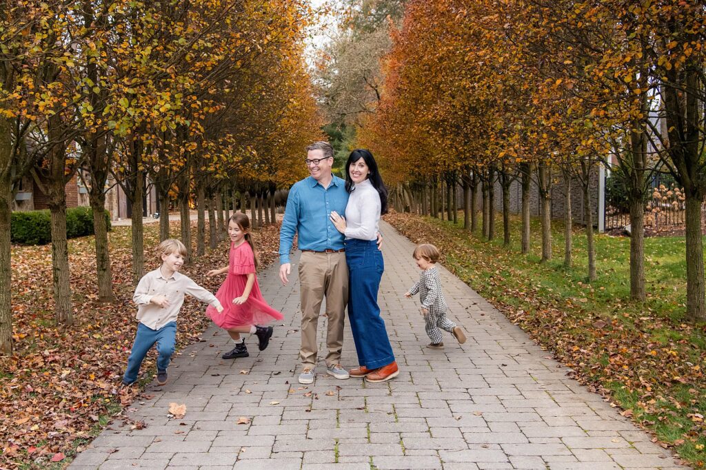 A family of two adults and three children stand and play on a tree-lined path with autumn leaves scattered on the ground.