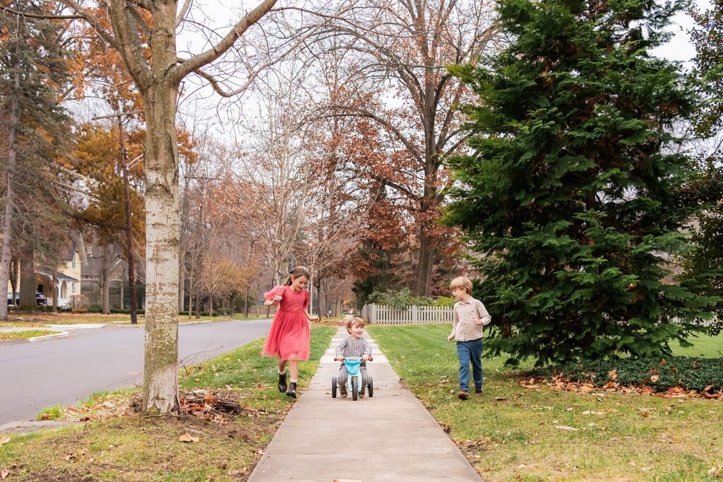 Three children play on a sidewalk; one rides a tricycle while the other two run beside him. Trees with sparse autumn leaves line the street and yard.
