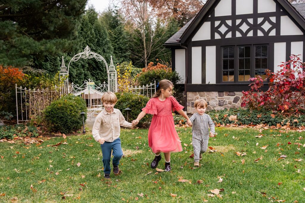Three young children hold hands and run on the grass in front of a house with Tudor-style architecture and a decorative garden gate.