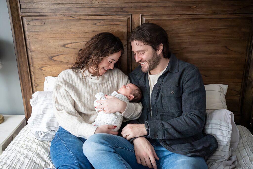 A man and woman sit on a bed, smiling and holding a newborn baby wrapped in a blanket.
