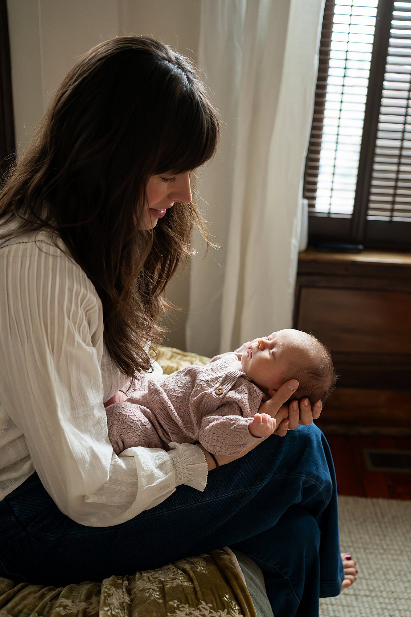 A woman with long brown hair sits on a bed holding a sleeping baby in her arms near a window with wooden blinds.