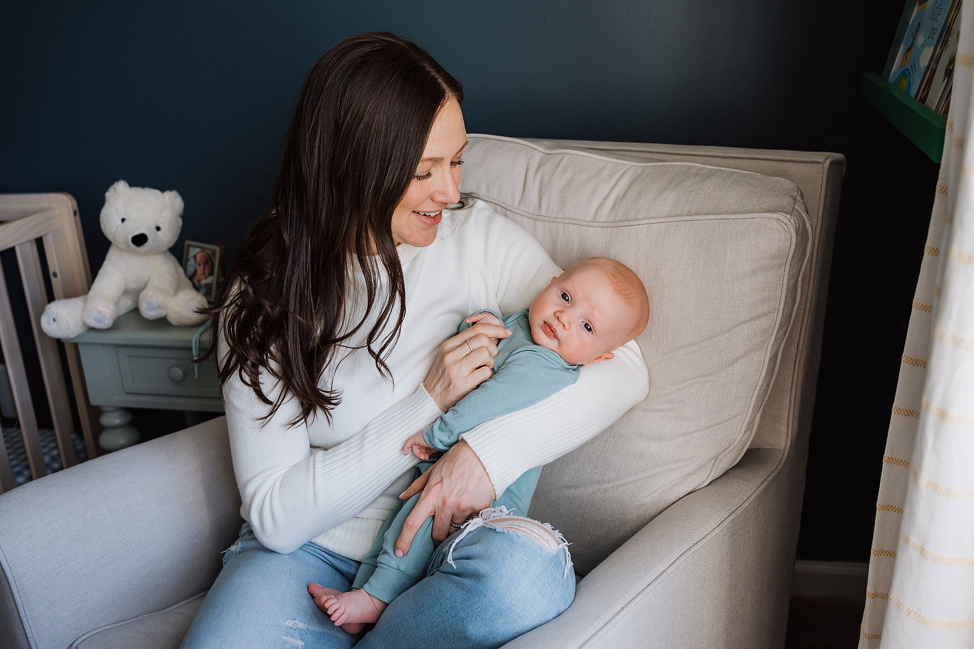 Woman sitting in an armchair holding a baby dressed in blue; a stuffed bear and framed photo are on a small table in the background.