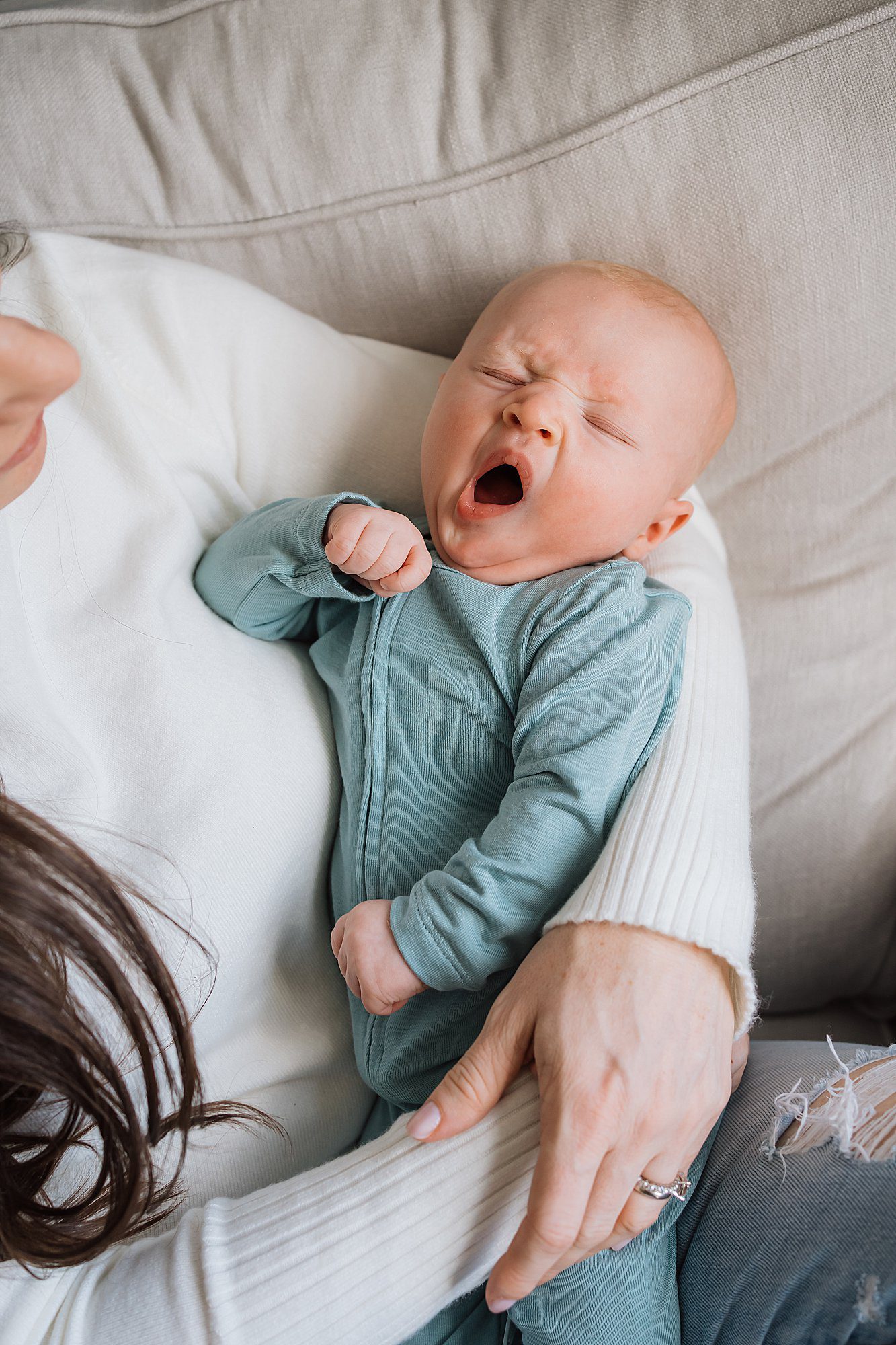 A baby in a teal onesie yawns while being held by an adult in a white sweater on a beige couch.