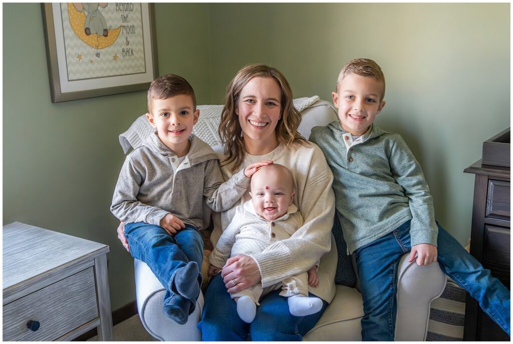 A woman sits in an armchair holding a baby on her lap, with two young boys sitting beside her, all smiling at the camera in a cozy room.