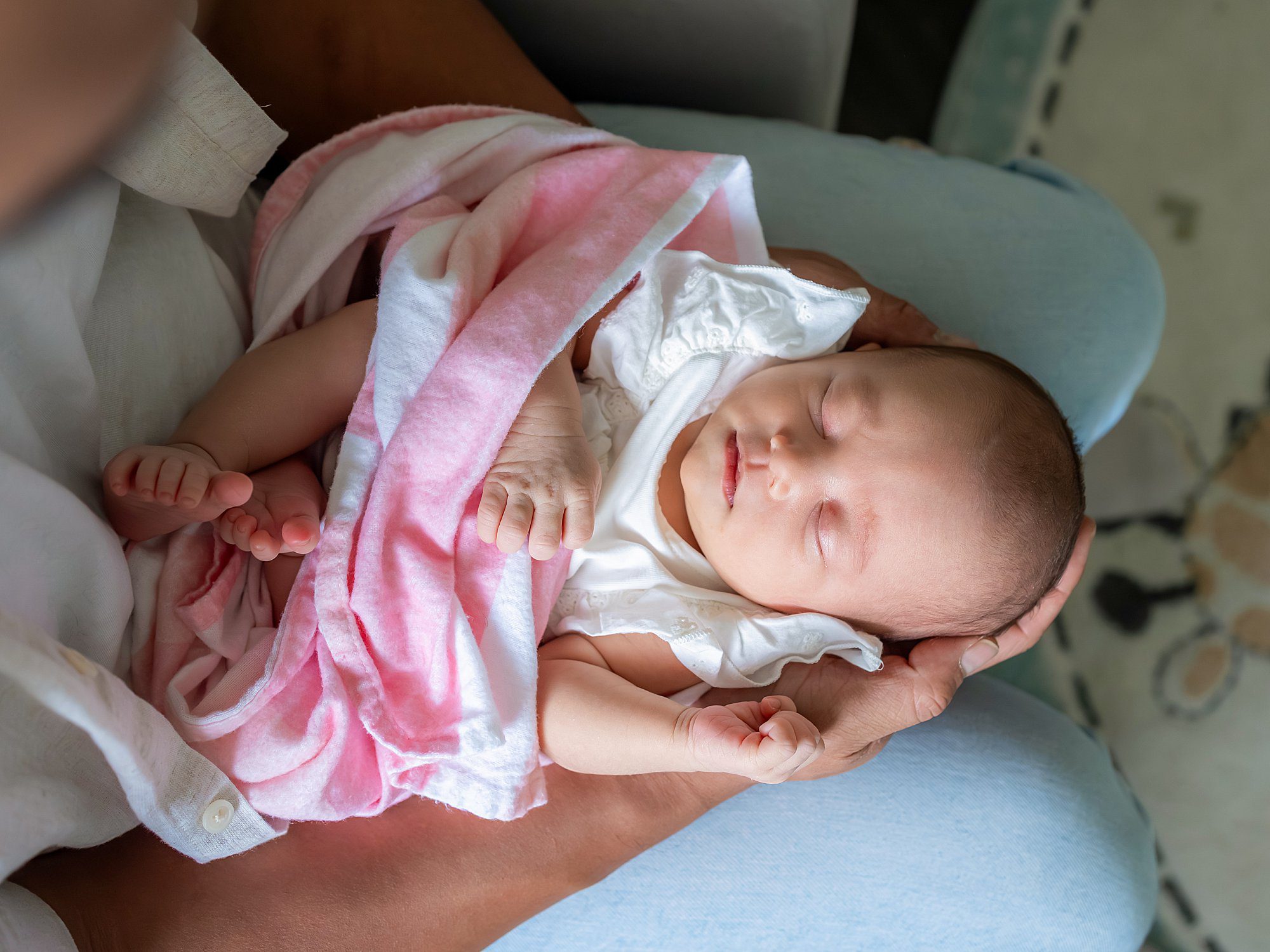 A newborn baby sleeps wrapped in a pink and white blanket, cradled in an adult's arms.