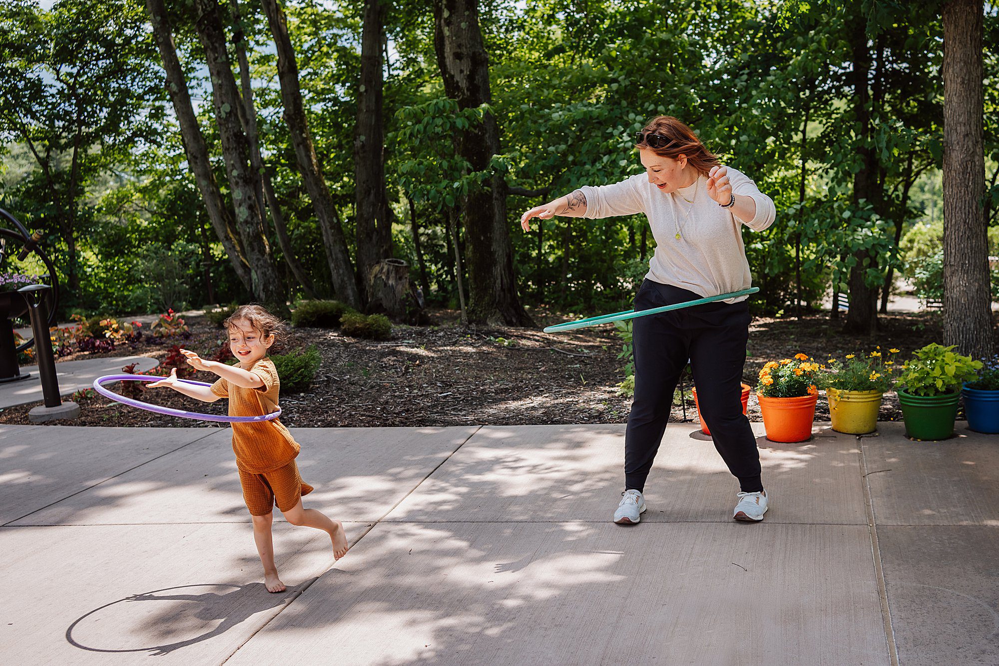 An adult and a child are outside on a sunny day, each spinning a hula hoop around their waist on a concrete patio with colorful potted plants nearby.