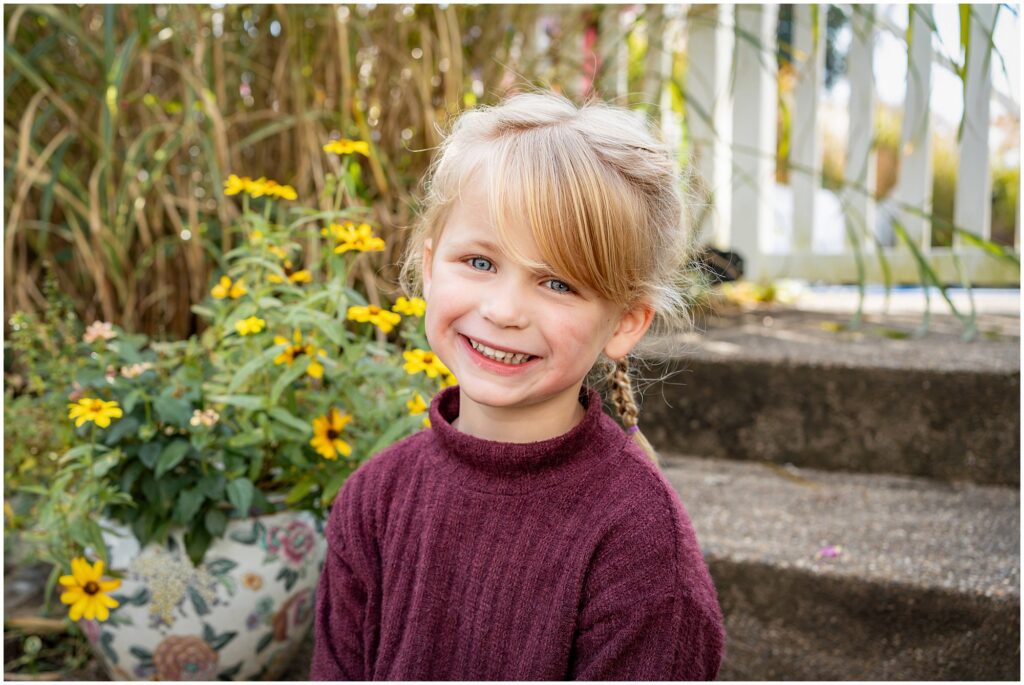 A young girl with blonde hair in braids, wearing a maroon sweater, sits outside on concrete steps next to a pot of yellow flowers.