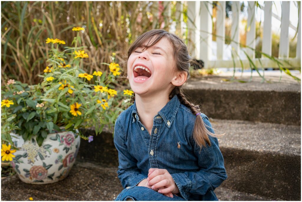 A young girl in a denim dress sits on outdoor steps, laughing, with yellow flowers in a patterned pot beside her.