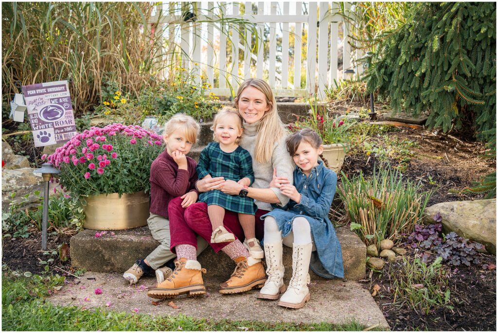 A woman sits outdoors on a stone step with three young children, surrounded by flowers and greenery, all smiling at the camera.