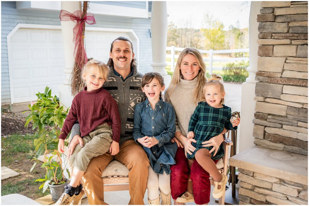 A family of five sits on a porch swing, smiling at the camera. There are two adults and three young children, with plants and a stone pillar in the background.