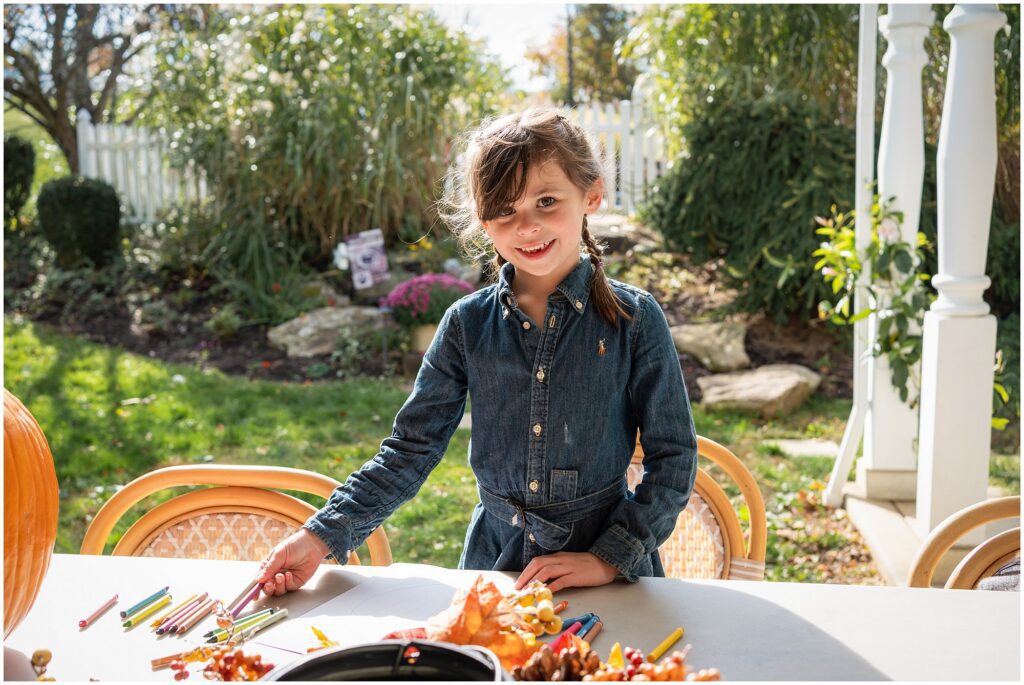 A young girl in a denim dress stands at an outdoor table with colored pencils, leaves, and a pumpkin, smiling at the camera on a sunny day.