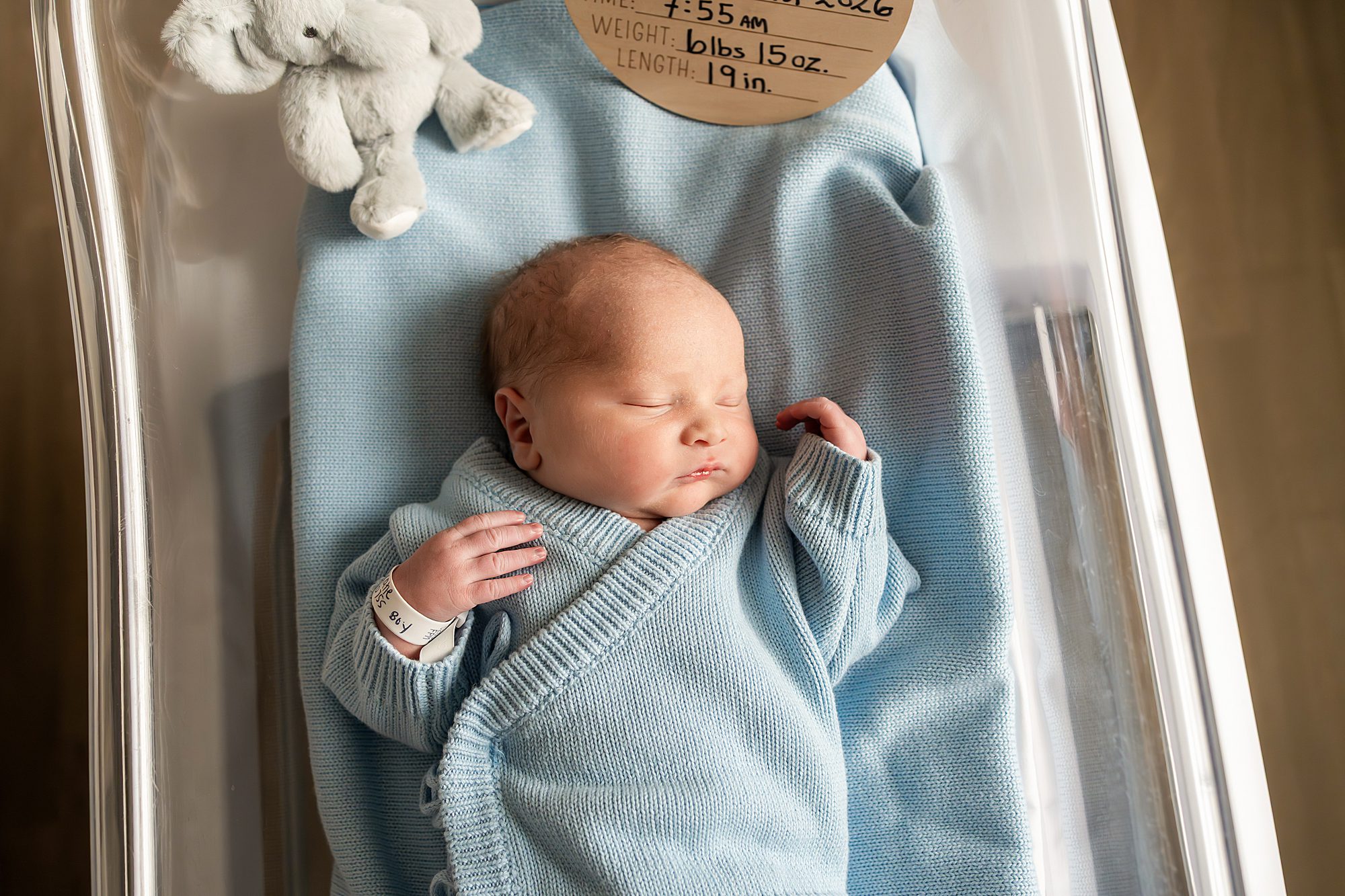 Newborn baby in a hospital bassinet, wrapped in a light blue blanket, with a hospital wristband and a small stuffed toy nearby.