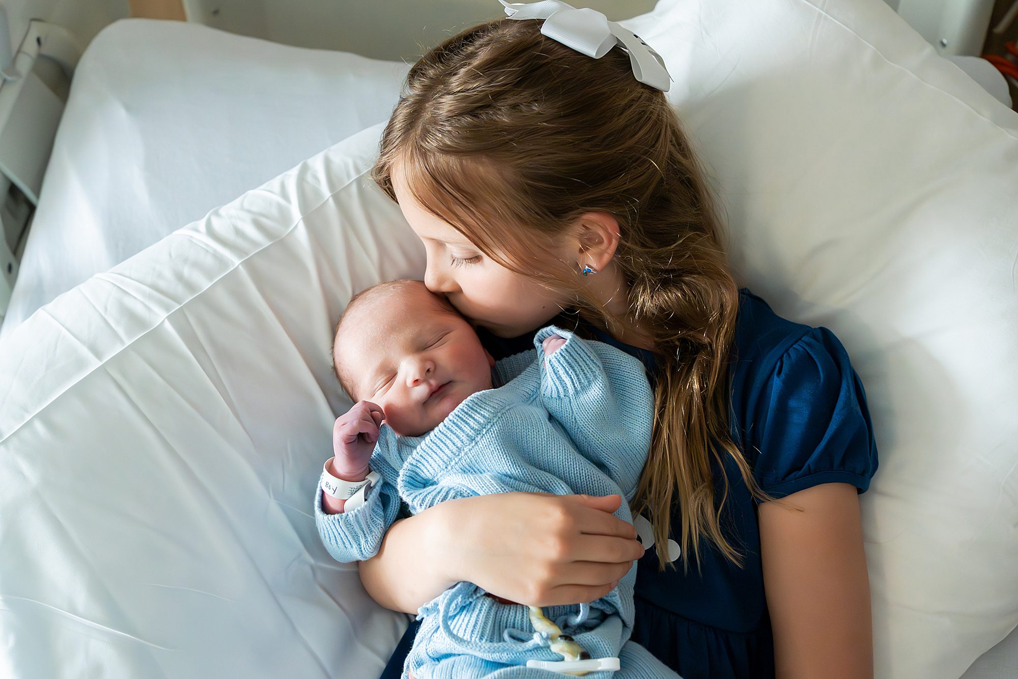 A girl in a blue dress sits on a bed, gently holding and kissing a newborn baby wrapped in a blue blanket.