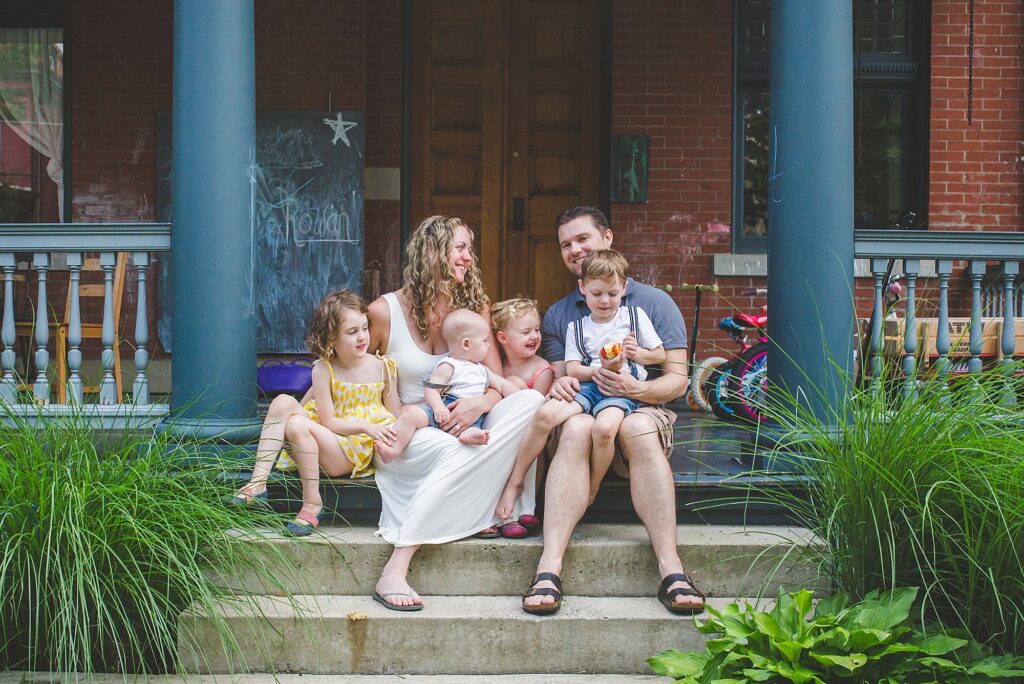 A family of two adults and four young children sit together on the steps of a porch in front of a brick house, surrounded by green plants.