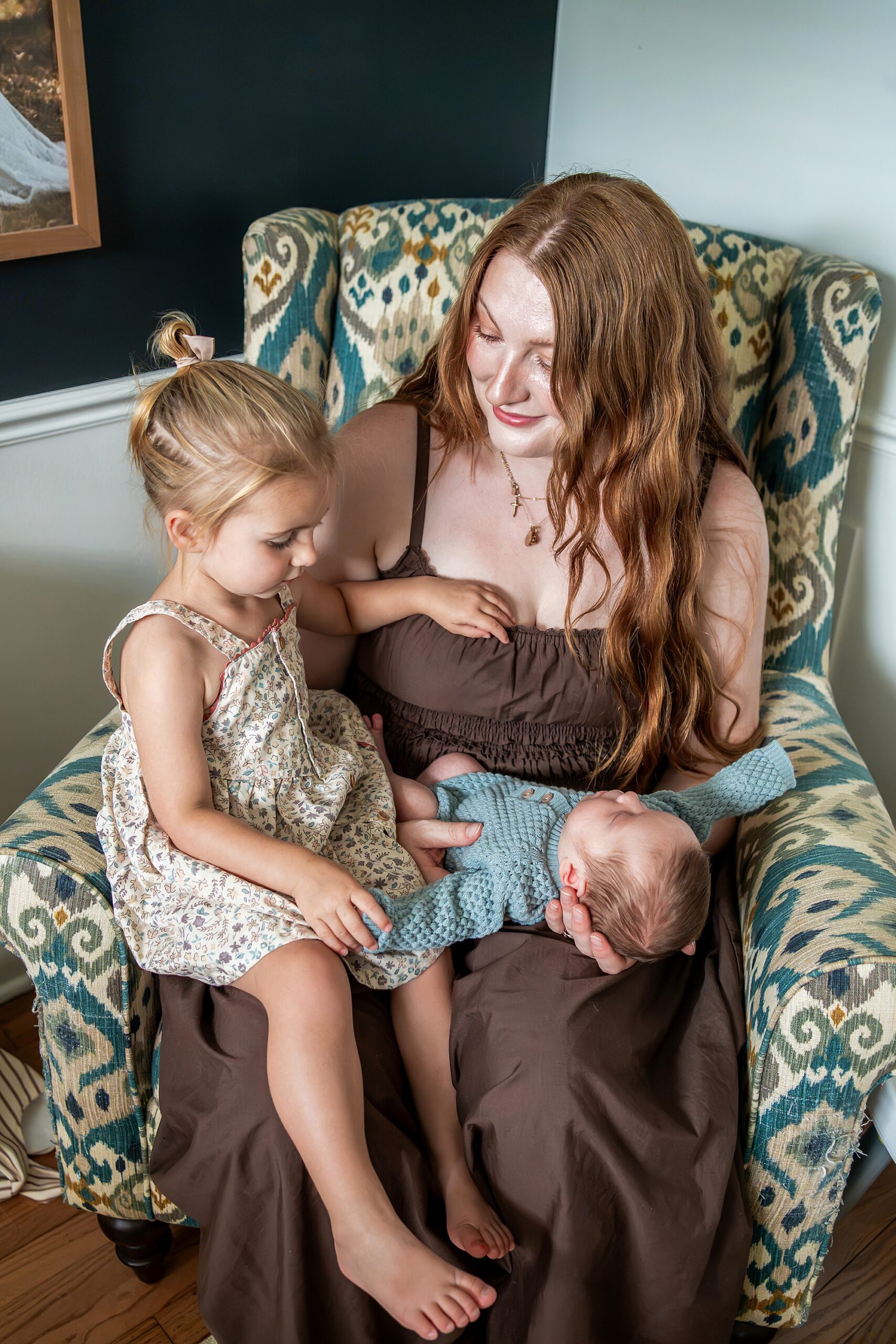 A woman sitting in a patterned armchair holds a newborn baby while a young girl sits beside her, touching the baby’s hand.