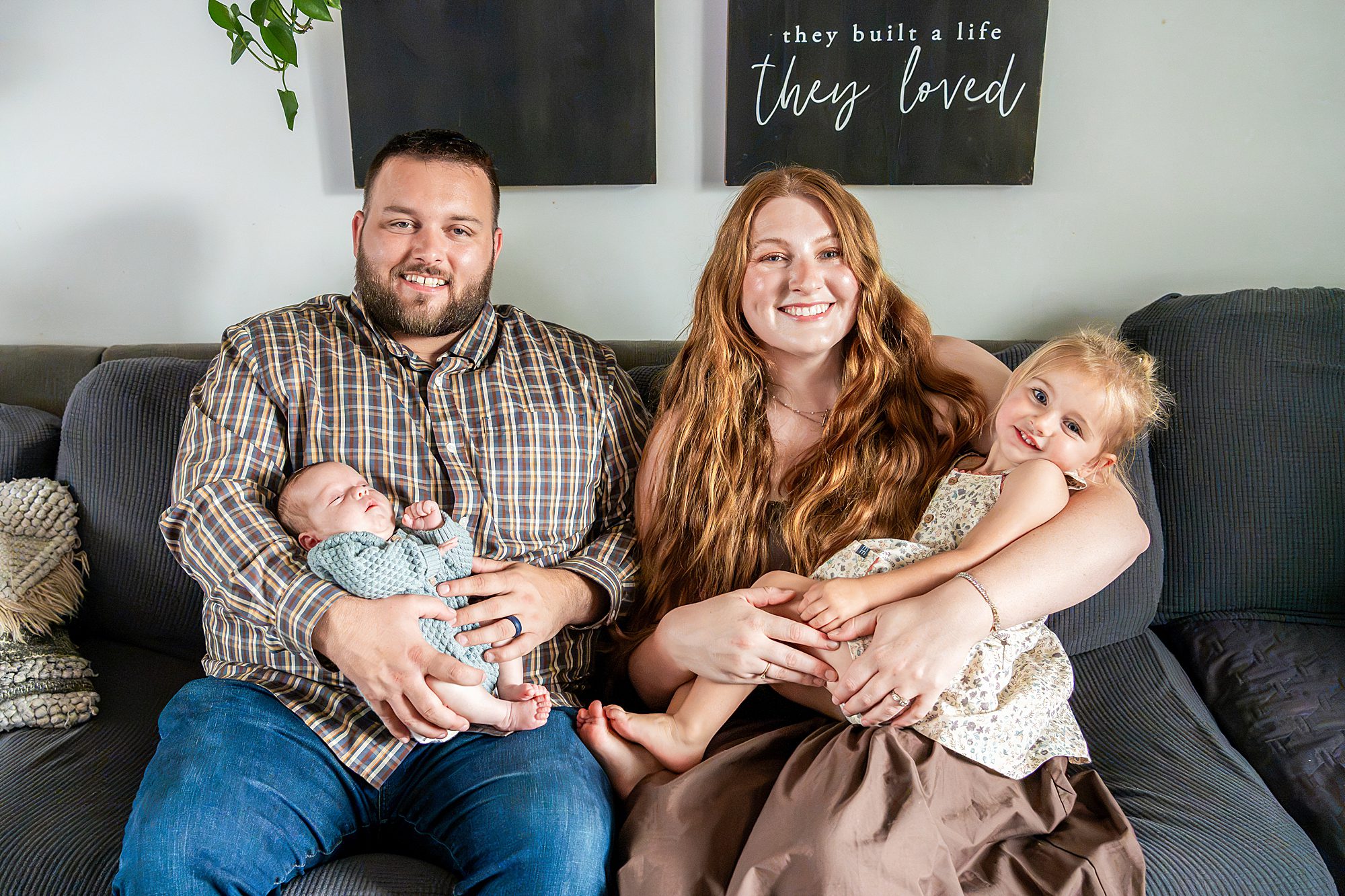 A family of four sitting on a sofa: a father holding a newborn, and a mother holding a young daughter.