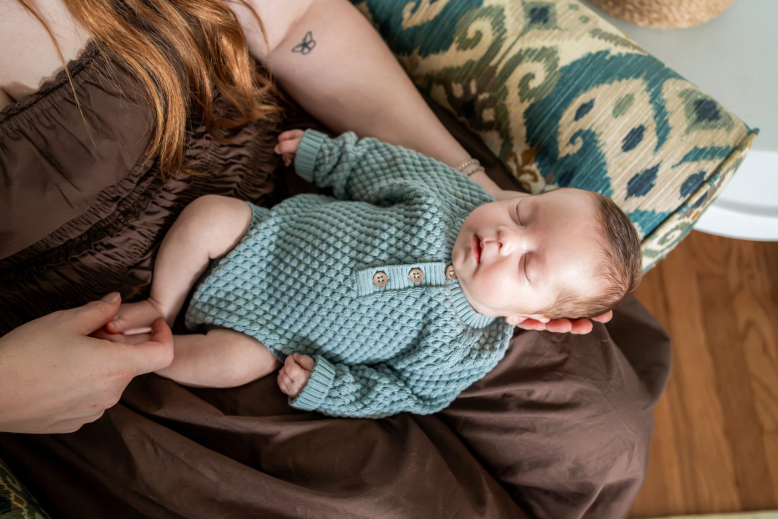A sleeping newborn baby in a sage green knit onesie held in a person’s lap.